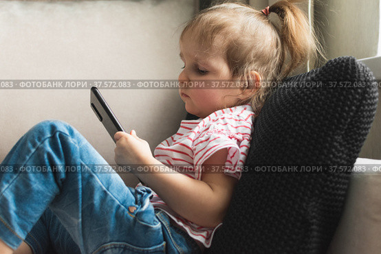 Little girl is sitting on sofa with smart phone. She's watching intently in it. Стоковое фото, фотограф Алдошина Екатерина / Фотобанк Лори