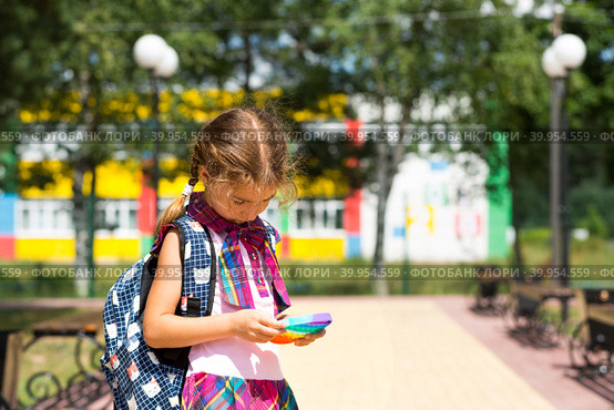 Little girl with a backpack and in a school uniform in the school yard plays pop it toy. Back to school, September 1. The pupil relaxes after lessons. Primary education, elementary class for student. Стоковое фото, фотограф Ольга Симонова / Фотобанк Лори