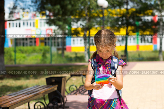 Little girl with a backpack and in a school uniform in the school yard plays pop it toy. Back to school, September 1. The pupil relaxes after lessons. Primary education, elementary class for student. Стоковое фото, фотограф Ольга Симонова / Фотобанк Лори