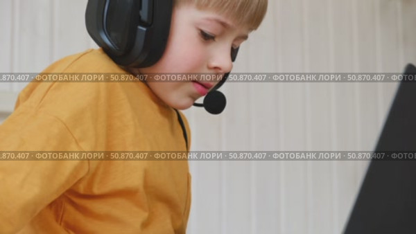 Little smiling 6 years old boy using a laptop with headphones. Child having video call with friends, granny at home in bed. Kid looking at display and waving hand. Childhood leisure activity concept. Стоковое видео, видеограф Марина Демидюк / Фотобанк Лори