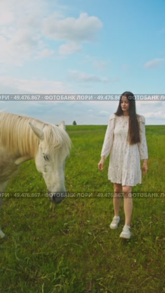 Lovely young girl is offering white horse some grass from her hand, standing in midst of a verdant meadow in the summer. Vertical. Стоковое видео, видеограф Strunov David / Фотобанк Лори