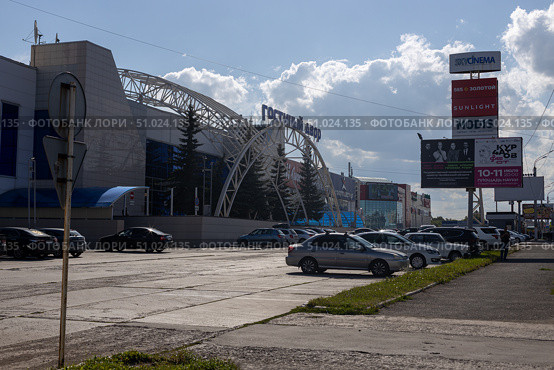 Magnitogorsk, Russian Federation - August 05, 2025: Gostiny Dvor shopping center entrance with modern architecture and blue glass facade, surrounded by trees and parked cars, showcasing urban lifestyle. Редакционное фото, фотограф Ирина Ручушкина / Фотобанк Лори