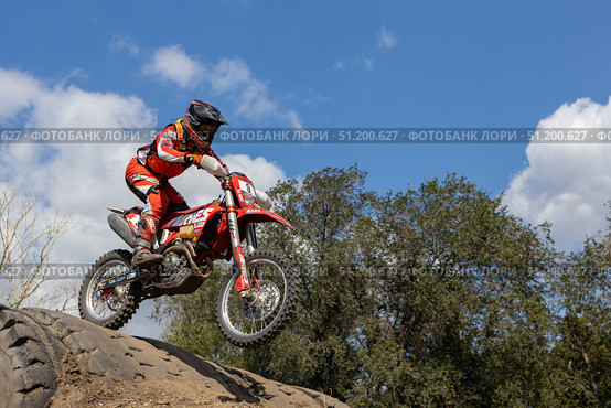 Magnitogorsk, Russian Federation - August 23, 2025: Male motocross rider in red gear jumps over dirt mound during enduro competition, showcasing skill and adrenaline in outdoor setting. Редакционное фото, фотограф Ирина Ручушкина / Фотобанк Лори