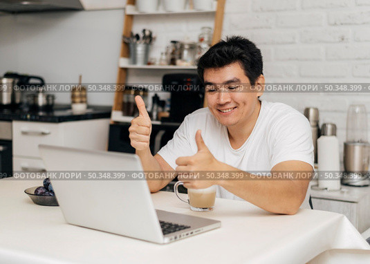 man home during pandemic showing thumbs up during video call laptop. Стоковое фото, агентство Ingram Publishing / Фотобанк Лори