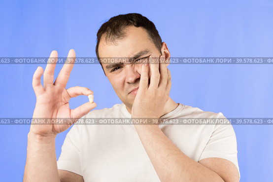 Man in a white T-shirt on a blue background holds a wisdom tooth in his hands after surgical tooth extraction.Man after an operation to remove wisdom teeth.Pain in wisdom teeth, concept of dentistry. Стоковое фото, фотограф Регина Ерофеева / Фотобанк Лори