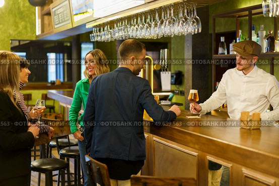 Man ordering beer from barkeeper while standing near happy talking women on weekend day in pub. Clients and bartender in an elegant bar. Стоковое фото, агентство Ingram Publishing / Фотобанк Лори