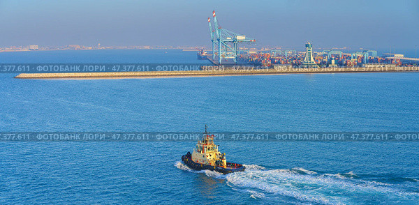 Manama, Bahrain - Jan 08 2024, Panoramic view of passing tugboat, a boat, in the background a commercial international port with cranes, pier, buildings and containers, in the daytime, Manama, Bahrain. Стоковое фото, фотограф Сергей Богомяко / Фотобанк Лори