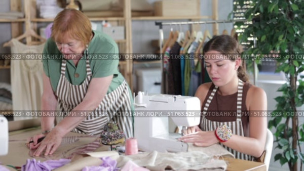 Mature dressmaker draws patterns of clothes on paper using curved rulers while an assistant seamstress works on a sewing machine. Стоковое видео, видеограф Яков Филимонов / Фотобанк Лори