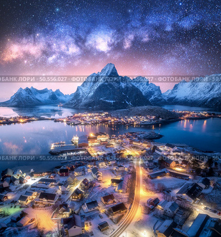 Milky Way over the snowy village, islands, rorbuer, city lights, sea, mountain, purple starry sky at winter night. Landscape with town, street illumination. Reine, Lofoten islands, Norway. Aerial view. Стоковое фото, агентство Ingram Publishing / Фотобанк Лори
