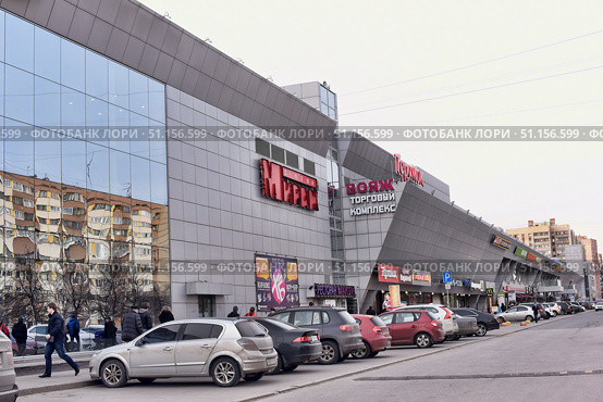 Modern shopping mall "Voyage" in the urban development. The building is made in a modern architectural style with cladding made of gray panels and large mirrored windows, which reflect residential buildings. On the facade there are signs of the cinema "Mireille", restaurants, shops and chain brands, among which McDonald's is visible. In front of the shopping center there is a parking lot with parked cars and pedestrians walking along the building. The scene conveys the atmosphere of everyday city life, the convenience of shopping malls and the dynamics of the metropolis. (2015 год). Редакционное фото, фотограф Анна Кривицкая / Фотобанк Лори