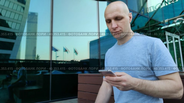 Молодой человек с мобильным телефоном сидит на скамейке возле зеркального небоскреба. Симпатичный парень со смартфоном. Красивый мужчина крупным планом. Общение, просмотр фото и видео, игры, интернет. A young man with a mobile phone is sitting on a bench near a mirrored skyscraper. Cute guy with a smartphone. A handsome man in close-up. Communication, viewing photos and videos, games, Internet. Стоковое видео, видеограф Ирина / Фотобанк Лори