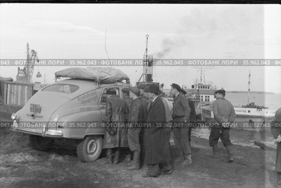 Молодые люди заглядывают в автомобиль марки Победа, они находятся на берегу водоёма, стрела погрузочного крана и речной паром на заднем плане, поездка Владивосток-Кронштадт, 1958. Редакционное фото, фотограф Варфоломей Тетерин / Фотобанк Лори