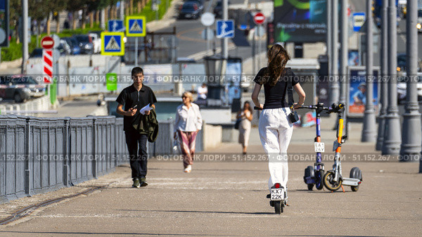 Moscow July 25 2025, People walking on the sidewalk in the city, a girl in white pants and a black T-shirt is fast riding a scooter on the sidewalk bypassing pedestrians. Редакционное фото, фотограф Скрипачев Антон / Фотобанк Лори