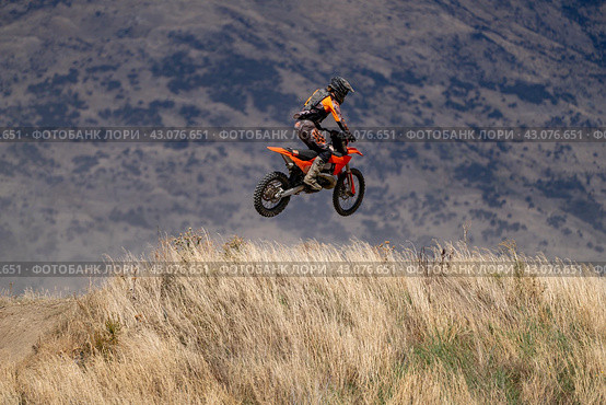 Motocross rider in orange and black gear performing a mid-air jump over a hill in Wanaka, New Zealand, against a mountainous backdrop. (2024 год). Редакционное фото, фотограф Евгений Дробитько / Фотобанк Лори