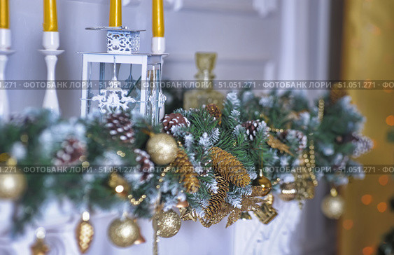 New Year's composition for interior decoration. On the white mantelpiece there is a garland of artificial pine with snow-covered branches, cones, golden balls and decorative elements. The decorations are complemented by golden cones, beads and figurines, creating an atmosphere of luxury and celebration. In the background you can see a white metal lantern with openwork patterns and yellow candles in tall white candlesticks. Soft bokeh in the background gives the composition coziness and warmth, and golden accents harmoniously combine with green and white colors. Стоковое фото, фотограф Анна Кривицкая / Фотобанк Лори