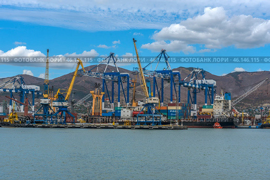 Novorossiysk Commercial Sea Port, one of the largest ports in Russia, located on the Black Sea coast. The frame shows cargo ships, port cranes, container yards and part of the industrial infrastructure. In the background are the mountain ranges surrounding the city.Numerous blue and yellow gantry cranes used for loading and unloading ships.Cargo containers stored in the port area.Commercial ships at the berths undergoing cargo operations.Tugboats used to maneuver large ships.Mountains, characteristic of the Novorossiysk landscape.Bright blue sky with clouds, creating a contrast with the industrial landscape. (2023 год). Редакционное фото, фотограф Анна Кривицкая / Фотобанк Лори