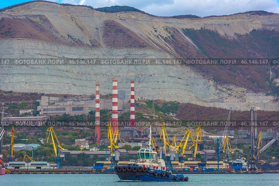 Novorossiysk port against the backdrop of the famous Markotkh ridge. In the foreground is a tugboat, and in the center are three red-and-white chimneys of the Novorossiysk cement plant, one of the oldest enterprises in the region. Numerous port cranes and warehouses are located along the shore.A tugboat in the port waters.An industrial zone with cement plant chimneys.Yellow port cranes unloading ships.The mountains of the Markotkh ridge with their characteristic limestone layers.Industrial buildings and warehouse complexes.Dense vegetation on the mountain slopes. (2023 год). Редакционное фото, фотограф Анна Кривицкая / Фотобанк Лори