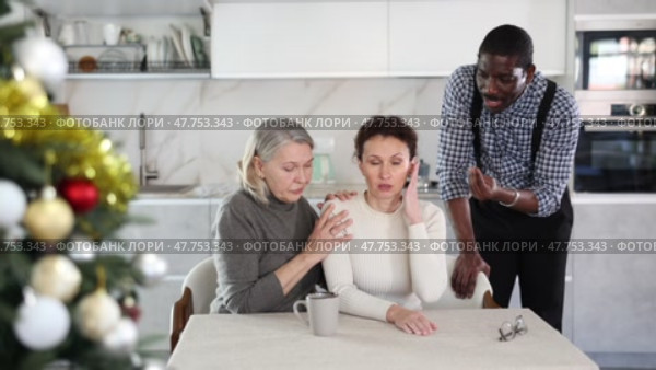 Old woman trying to calm angry middle-aged woman sitting at table while man quarreling to her standing behind in the kitchen decorated with Xmas-tree. Стоковое видео, видеограф Яков Филимонов / Фотобанк Лори
