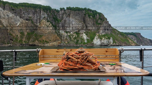 On the yacht, on the table, there is a tray. Boiled snow crabs are stacked in a slide. Legs and claws are visible. The background is the ocean, the rocky coast of Kamchatka. Стоковое фото, фотограф Вера Ивановна Кубарева / Фотобанк Лори