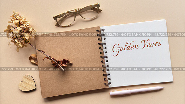 Open scrapbook with text Golden Years on a white page, surrounded by dried flowers, wooden heart, glasses, and pen on a beige background. Стоковое фото, фотограф Аnna Ivanova / Фотобанк Лори