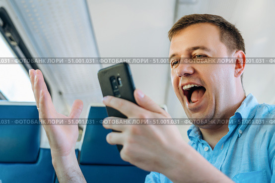 Overjoyed caucasian man looks at smartphone screen with mouth opened in train commuting to work. Excited man looking with wide open eyes into smartphone being shocked by what he sees on way to work. Стоковое фото, фотограф Данила Большаков / Фотобанк Лори