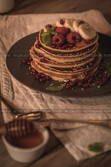 Pancakes with raspberries, banana slices, pomegranate seeds and honey on wooden vintage table. Стоковое фото, агентство Ingram Publishing / Фотобанк Лори