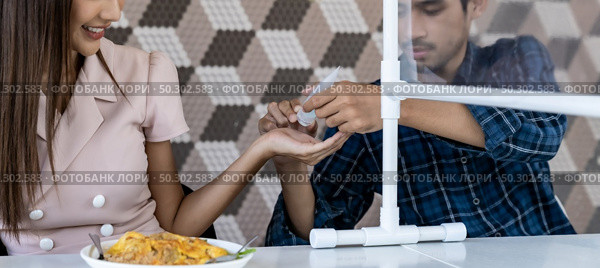 Panoramic asian man put alcohol gel hand sanitizer to his partner before eating at new normal social distance restaurant with table partition. Restaurant new normal lifestyle. Web banner crop. Стоковое фото, агентство Ingram Publishing / Фотобанк Лори