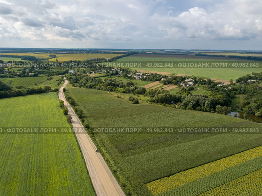 Panoramic view from drone to the countryside with a country buildings, dirt road and agricultural fields against cloudy sky in the summer at sunset.. Aerial view from a bird’s-eye view to a rural landscape with a village, dirt road and agricultural fields of planted crops. Стоковое фото, агентство Ingram Publishing / Фотобанк Лори