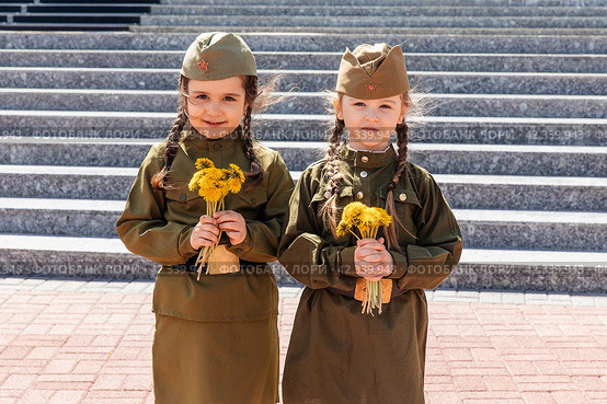 Patriotic education of children. Little girls with long braids in a military uniform, military cap, hold flowers in hands against the background of the memorial. Photo for the Victory Day on May 9. Стоковое фото, фотограф Ирина Михайличенко / Фотобанк Лори