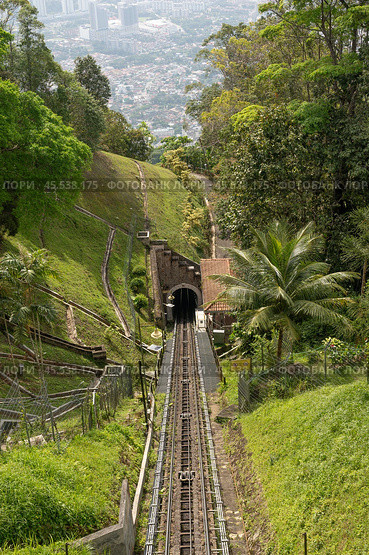 Penang Hill funicular railway in Malaysia, moving down hillside, surrounded by dense tropical forest. Offers panoramic views of lush greenery and city skyline in distance. must-visit spot for tourists. Стоковое фото, фотограф Павел Муравьев / Фотобанк Лори