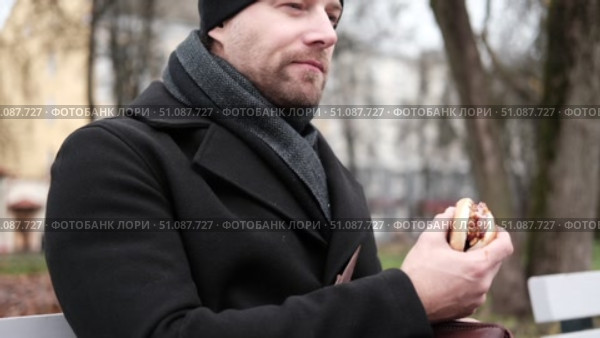 Pensive hipster drinking coffee sitting on a bench in the urban park. Handsome man wearing black coat eating street food, hot dog with paper cup in hand in the city. Stylish bearded middle-aged male. Стоковое видео, видеограф Марина Демидюк / Фотобанк Лори