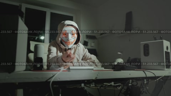 Person in hoodie and glowing mask sitting at desk in dark room, concept of hidden identity and digital privacy in online space. Perfect for cybersecurity, anonymity and internet themes. Стоковое видео, видеограф Енваев Андрей / Фотобанк Лори