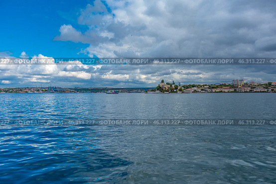 Picturesque seascape of Sevastopol Bay. In the foreground is a smooth water surface reflecting the sky and clouds. In the distance on the horizon is visible the coast with city buildings, green spaces and port facilities.On the right, on the hill, historical buildings are visible, probably one of the iconic objects of Sevastopol. Voluminous clouds hang over the city, creating an atmosphere of maritime climate, changeable weather and natural grandeur. The picture conveys the spirit of the sea city, its connection with history, navy and nature. Стоковое фото, фотограф Анна Кривицкая / Фотобанк Лори