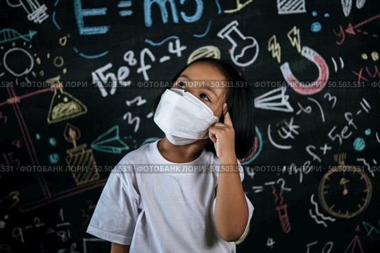 Portrait of a little girl wearing eyeglasses and facemask at school to avoid the spread of coronavirus, new normal with schoolchild,  blure blackboard in background. Стоковое фото, агентство Ingram Publishing / Фотобанк Лори