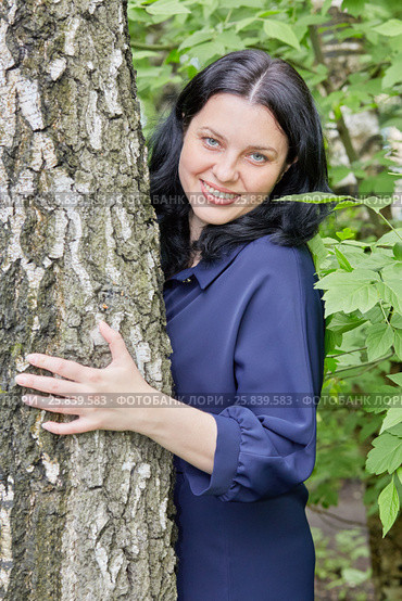 Portrait of smiling dark-haired woman peeking out from behind the tree in green summer park. Стоковое фото, фотограф Losevsky Pavel / Фотобанк Лори