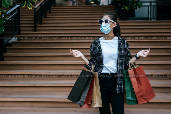 Portrait of young Asian woman in face mask wearing eyeglasses standing on stairs with shopping paper bag. she smile with happy, New normal Lifestyle and shopping concept. Стоковое фото, агентство Ingram Publishing / Фотобанк Лори