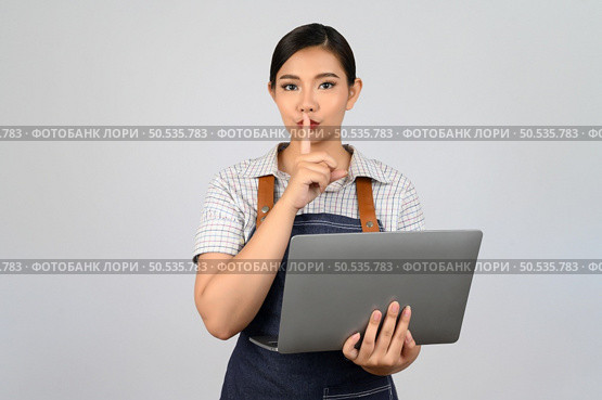 Portrait of young asian woman in waitress uniform holding a laptop computer and finger on mouth posture to make a quiet, copy space to insert your advertisement isolated on white background. Стоковое фото, агентство Ingram Publishing / Фотобанк Лори