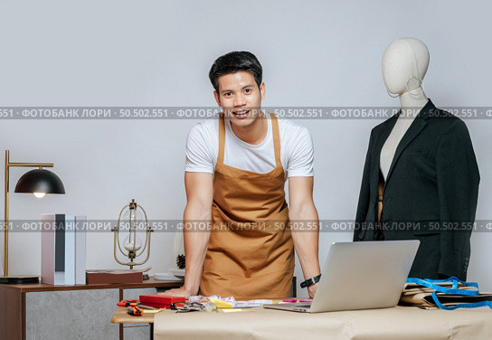 Portrait Young handsome male designer and laptop computer for searching good idea to design fashion clothing, mannequin and equipment with full of tailoring tools on desk in studio. Стоковое фото, агентство Ingram Publishing / Фотобанк Лори