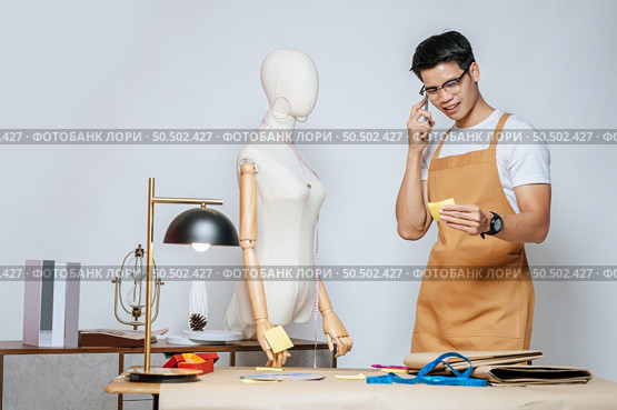 Portrait Young male fashion designer use smartphone during working with mannequin and take a note at the studio at home,  full of tailoring tools and equipment. Стоковое фото, агентство Ingram Publishing / Фотобанк Лори
