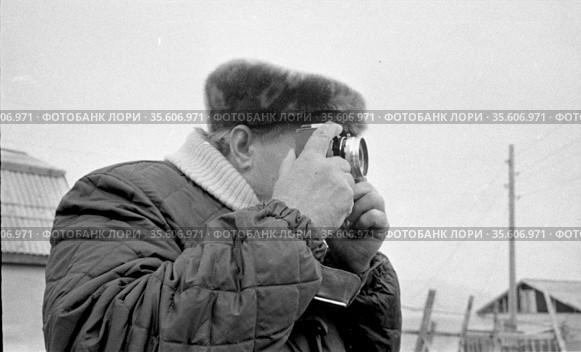 Портрет мужчина с фотоаппаратом, человек с камерой занимается фотографированием птицефермы, деревянные постройки и опора ЛЭП на заднем плане, Лорино, Чукотка, 1970. Редакционное фото, фотограф Варфоломей Тетерин / Фотобанк Лори