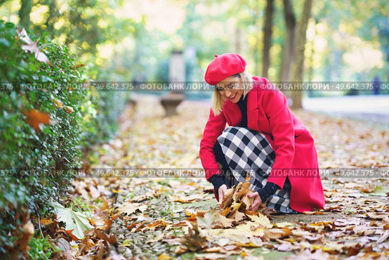 Positive female in checkered skirt and red outerwear picking fallen leaves from path in autumn park. Smiling woman collecting leaves from ground in park. Стоковое фото, агентство Ingram Publishing / Фотобанк Лори