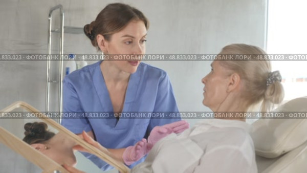 Positive interested mature woman looking at mirror while skilled female cosmetologist showing results of hardware facial procedure in medical esthetic office. Стоковое видео, видеограф Яков Филимонов / Фотобанк Лори