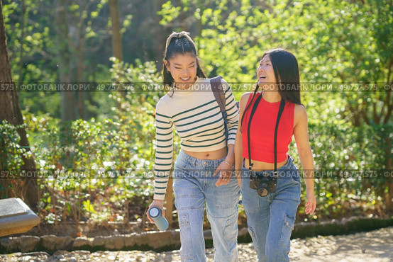 Positive young Asian female friends in casual outfits smiling and speaking about life while strolling together with thermos and camera in green forest of Alhambra. Cheerful Asian travelers walking in park and chatting. Стоковое фото, агентство Ingram Publishing / Фотобанк Лори