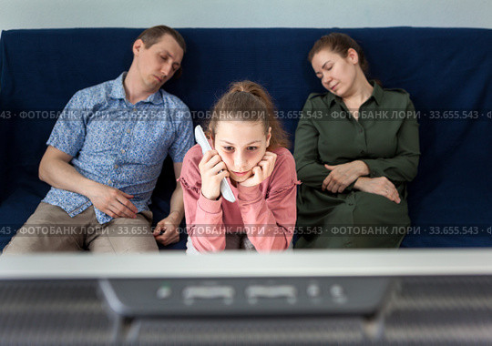 Pre-teen girl with sleepy look watching television set at night during her parents sleeping on couch, family at home. Стоковое фото, фотограф Кекяляйнен Андрей / Фотобанк Лори