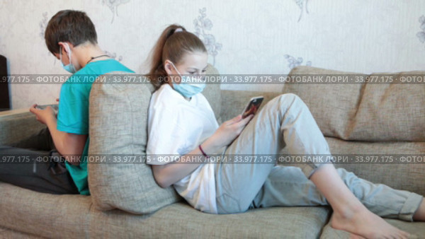 Preteen girl and boy playing online with cellphones while staying at home, children wearing face masks, sitting back to back. Стоковое видео, видеограф Кекяляйнен Андрей / Фотобанк Лори