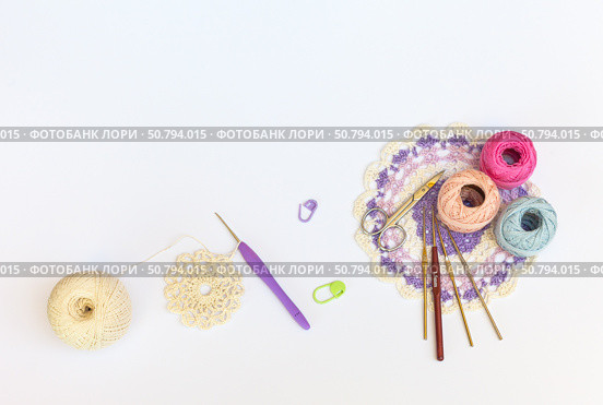 Process of crocheting an openwork round napkin with crochet hook. Set of hooks, skeins of cotton colored yarn, scissors and lace three-color doily on white background. Copy space, flat lay, close-up, top view. Стоковое фото, фотограф Виктория Катьянова / Фотобанк Лори