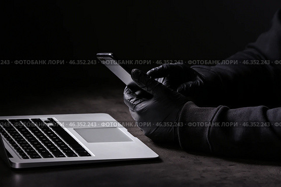 Professional hacker with laptop and mobile phone sitting at table on dark background. Стоковое фото, фотограф Pixel_shot / Фотобанк Лори