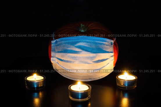 Pumpkin in medical disposable mask in dark and three lighted tea candles. Halloween and new normal concept. Стоковое фото, фотограф Зарифа Арсланова / Фотобанк Лори