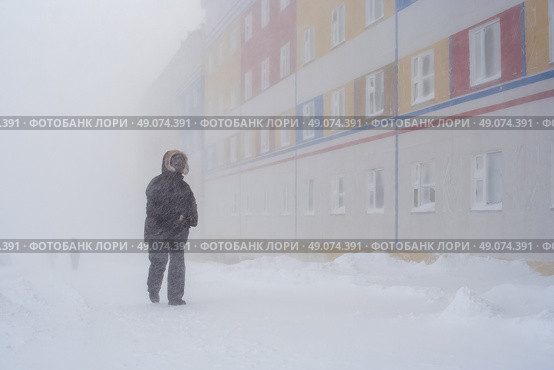 Пурга в городе. Мужчина идёт по заснеженной улице мимо разноцветного жилого дома. Метель с сильным ветром. Суровый полярный климат в Арктике. Холодная зимняя погода. Город Анадырь, Чукотский автономный округ (Чукотка), Дальний Восток России. Февраль. Стоковое фото, фотограф Андрей Степанов / Фотобанк Лори