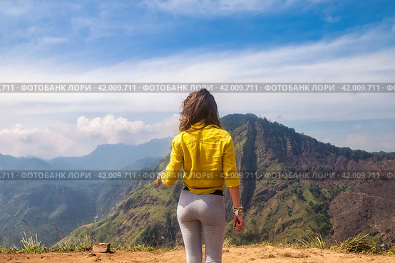 Rear view of lady enjoy at landscape Adam's peak mountain at tropical journey Sri Lanka, Ella, posing. From behind of adult woman in yellow jacket at landmark nature background. Copy ad text space. Стоковое фото, фотограф Aleksandr Golubev / Фотобанк Лори